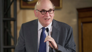 Paul Aisen is wearing a grey suit and holding a microphone at a podium